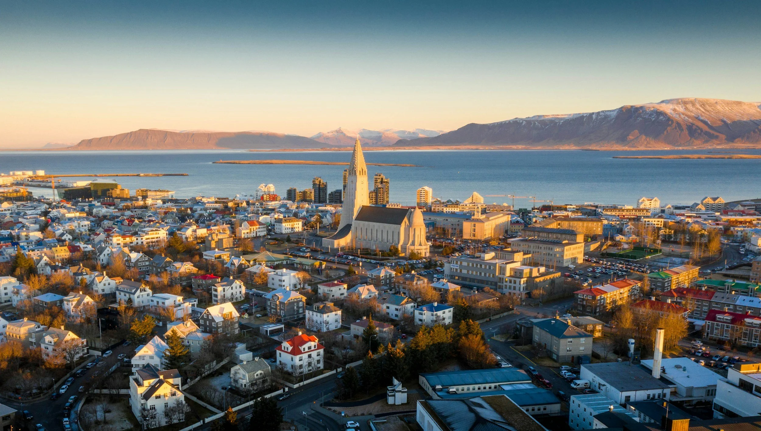 Aerial view of Reykjavik city center with a view of Hallgrimskirkja church, surrounded by residential and commercial buildings, with mountains and water in the background during sunset.