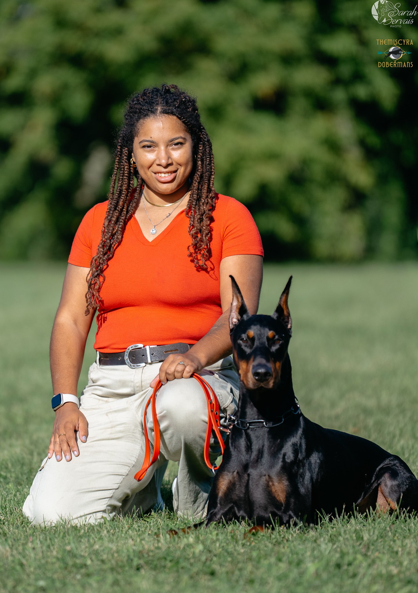 A woman dressed in an orange shirt and beige pants, kneeling on grass, with a black and tan Doberman dog sitting in front of her. The woman is smiling and holding the dog's orange leash. The background features blurred greenery.