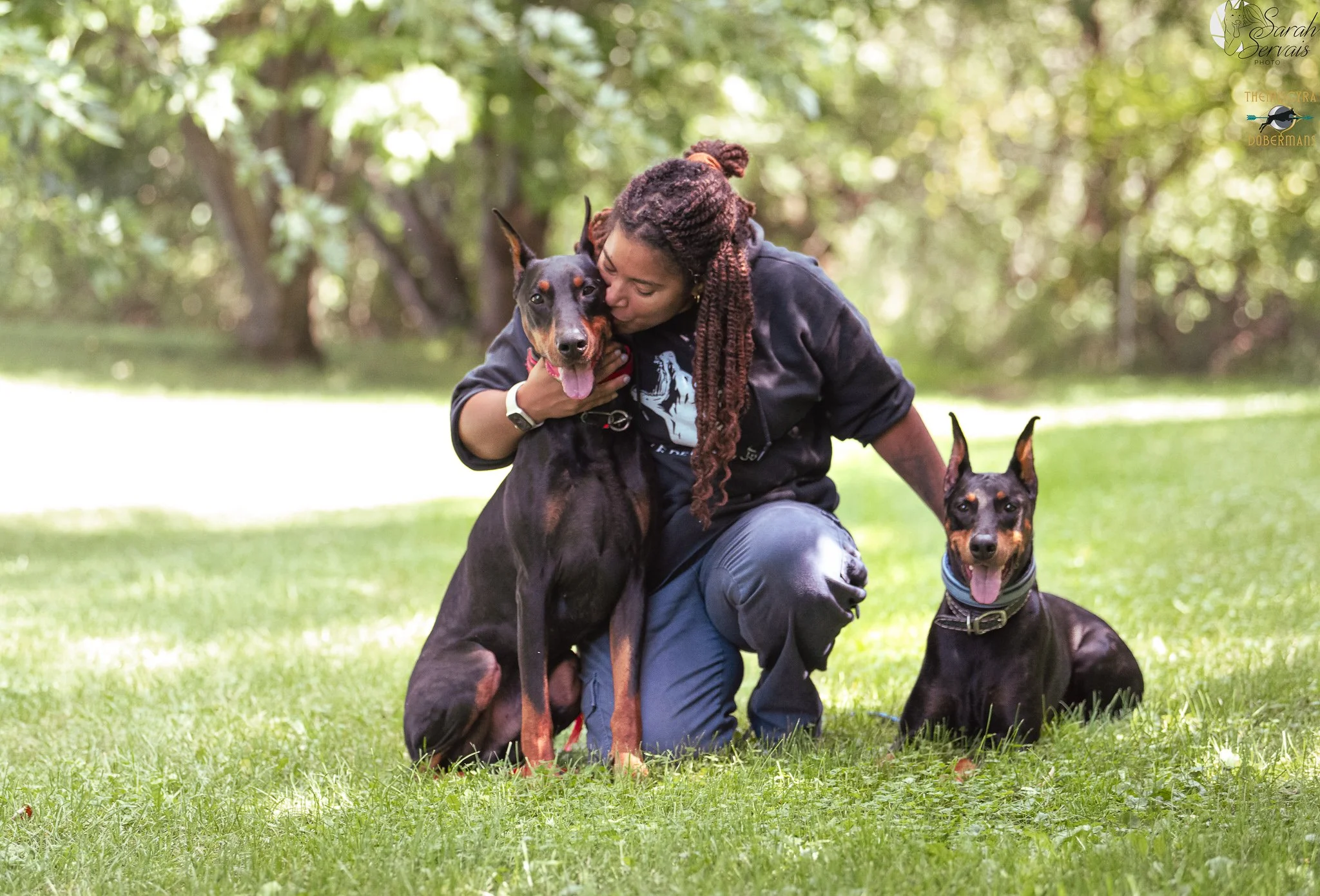 A woman with long braided twists hugging and kissing a large Doberman dog outdoors in a park, with another Doberman sitting nearby on grass, surrounded by trees.