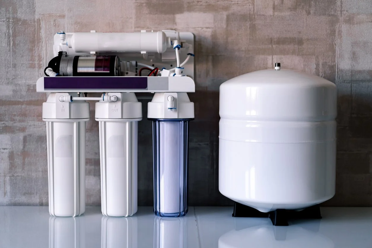 A water filtration system, including three filter housings and a storage tank, placed on a countertop with a gray textured wall in the background.