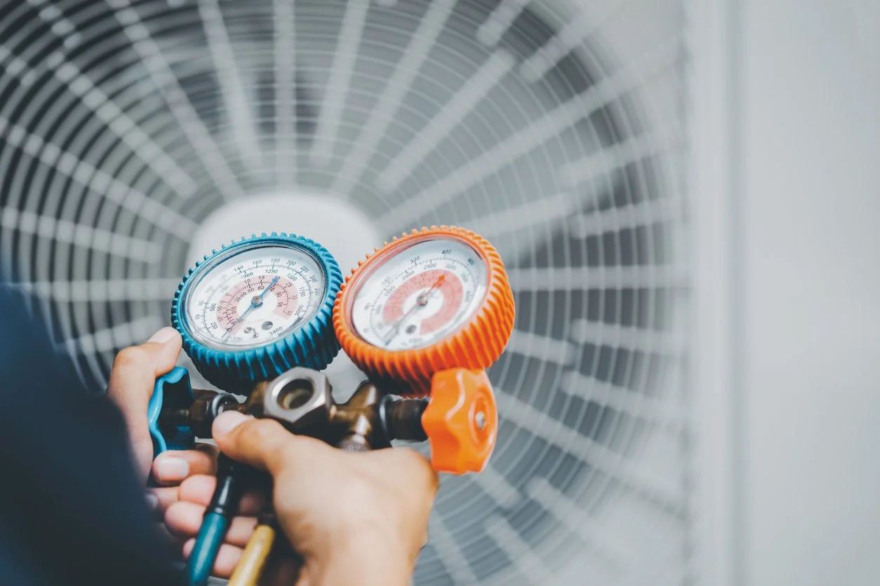 Close-up of a person's hands holding refrigeration service manifold gauges connected to an air conditioning unit, with an industrial fan in the background.