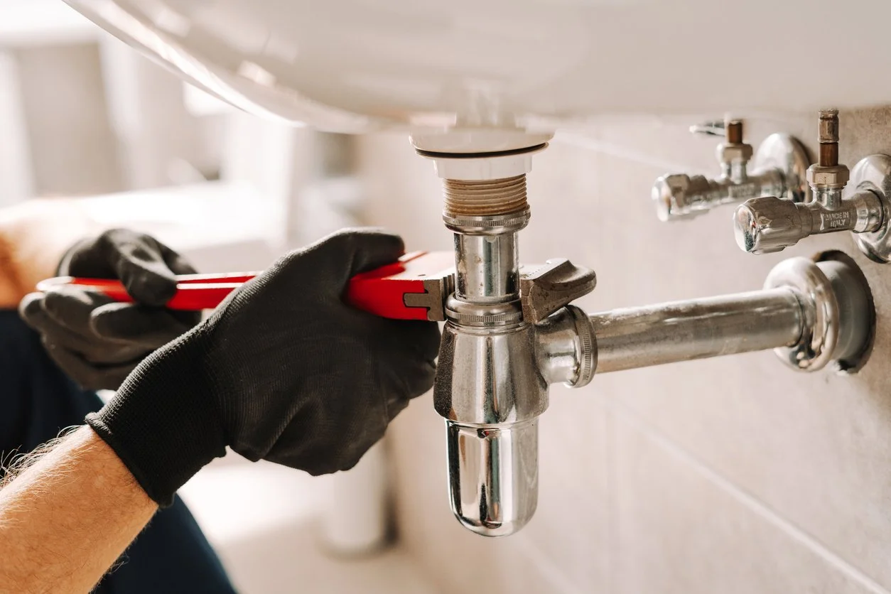 A person in black gloves is using a red adjustable wrench to tighten a pipe fitting under a sink.
