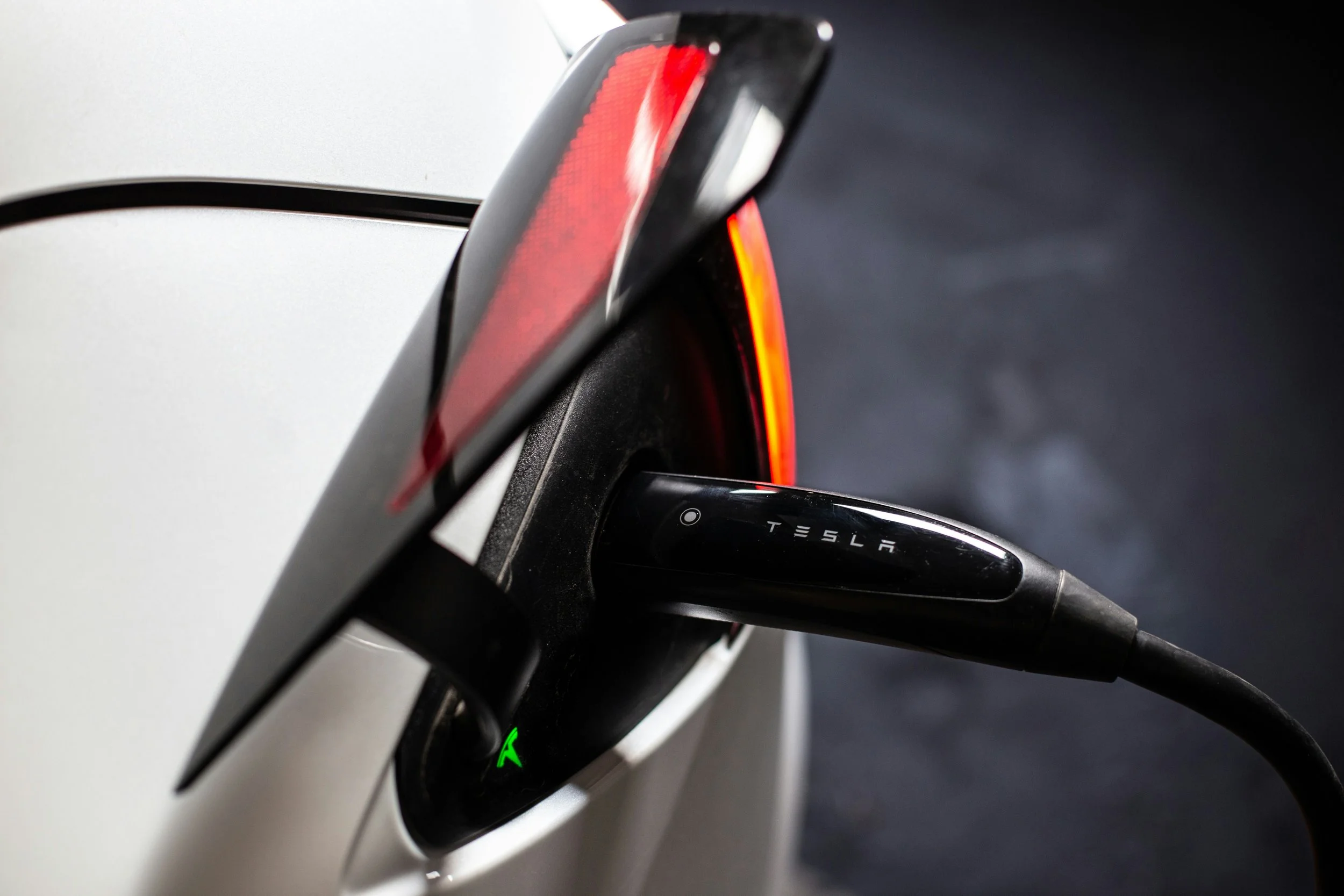 Close-up of a Tesla electric vehicle charging port with a charging cable plugged in, showing a red taillight and the Tesla logo on the connector.