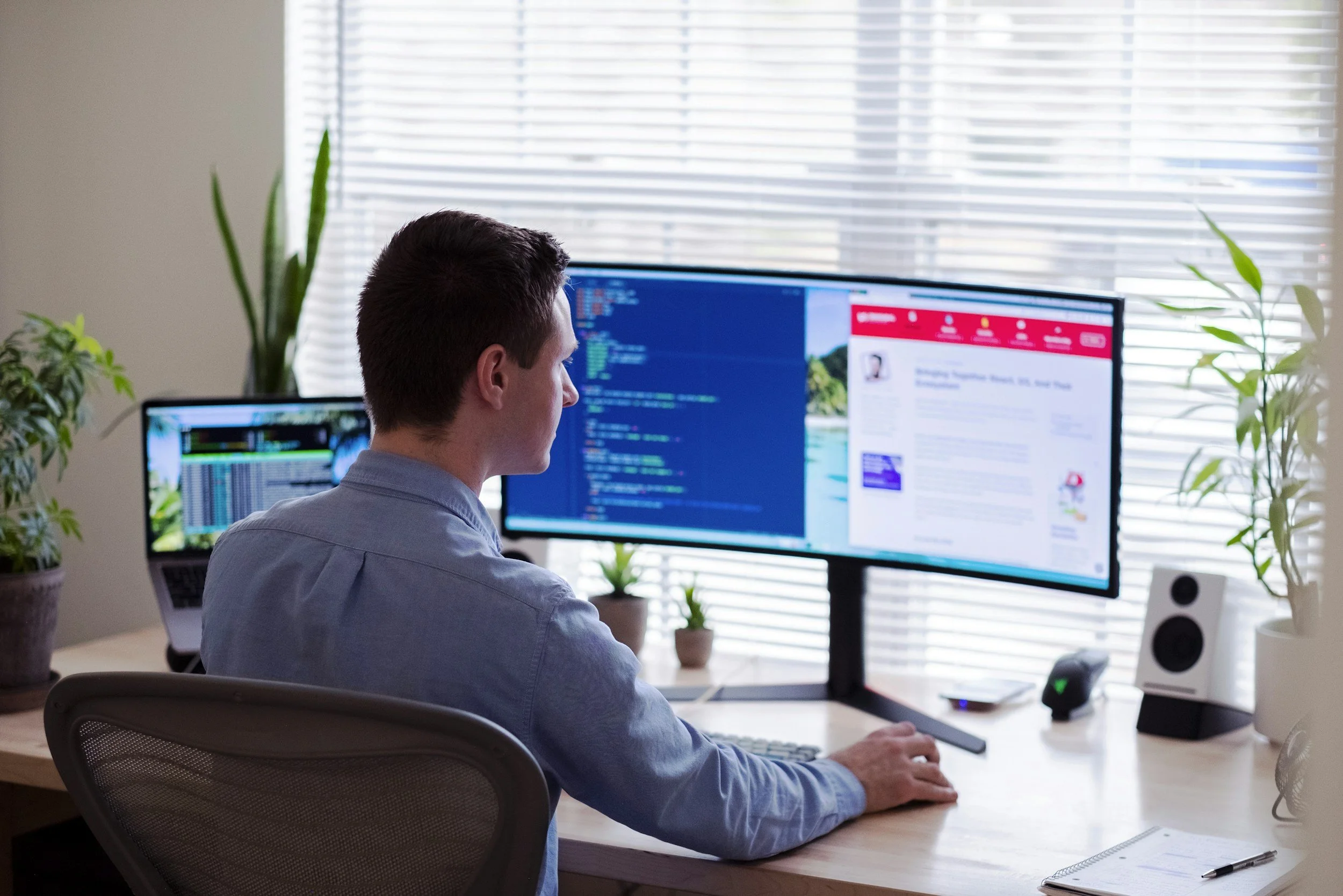A man working at a desk with two computer monitors, one displaying code and the other a webpage, in a bright room with large windows and potted plants.