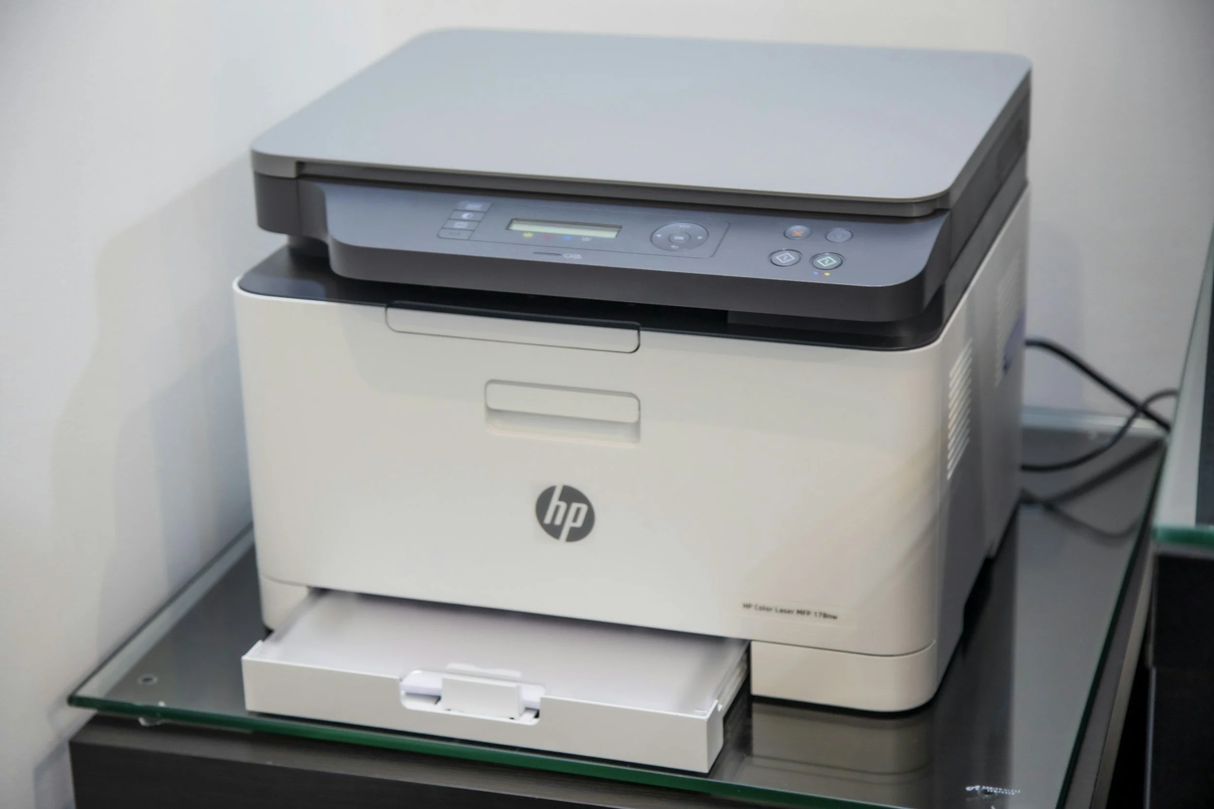 A white HP all-in-one laser printer on a black table against a white wall.