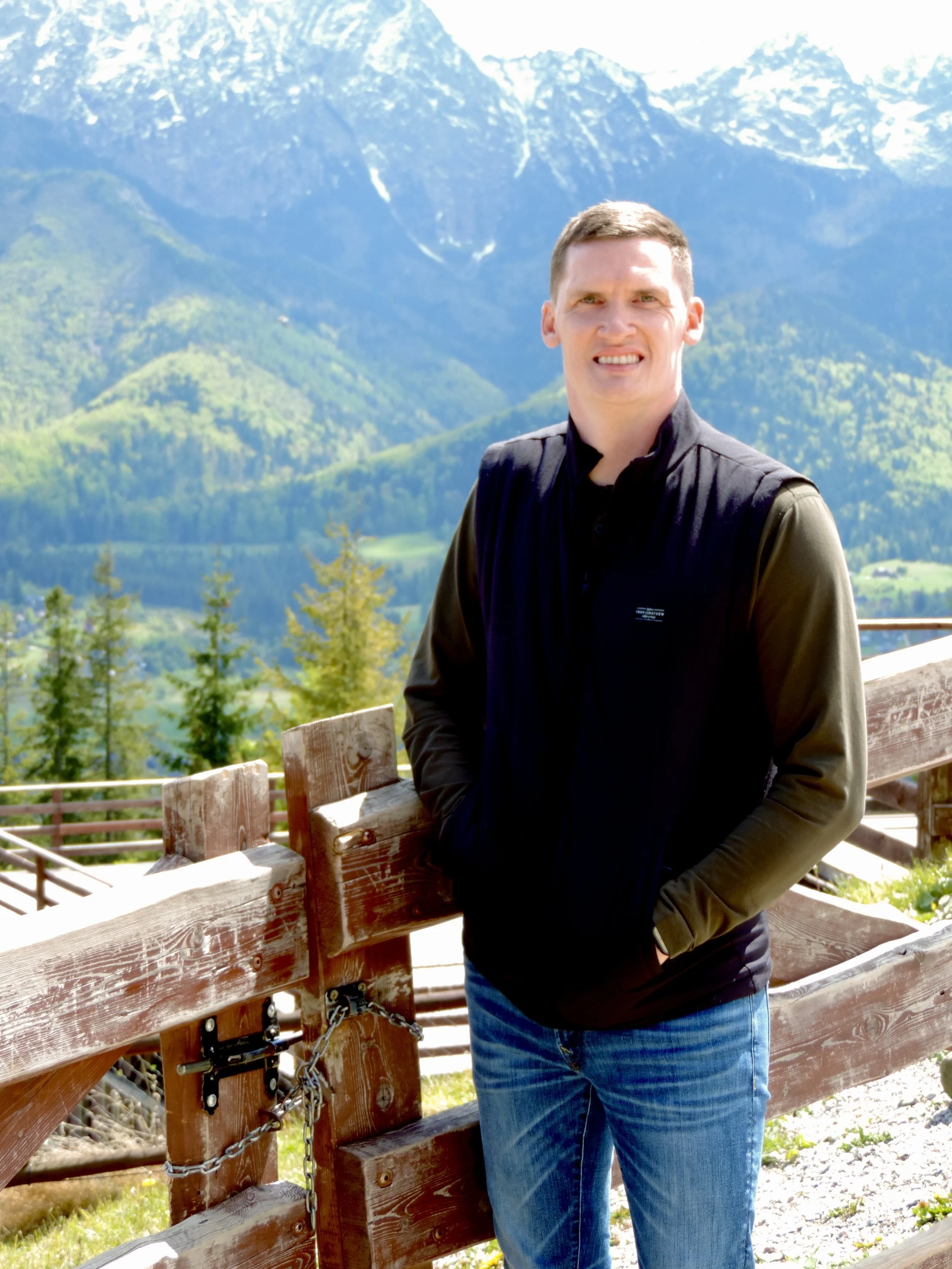 A man wearing a black jacket and blue jeans standing next to a wooden fence with mountains and greenery in the background.