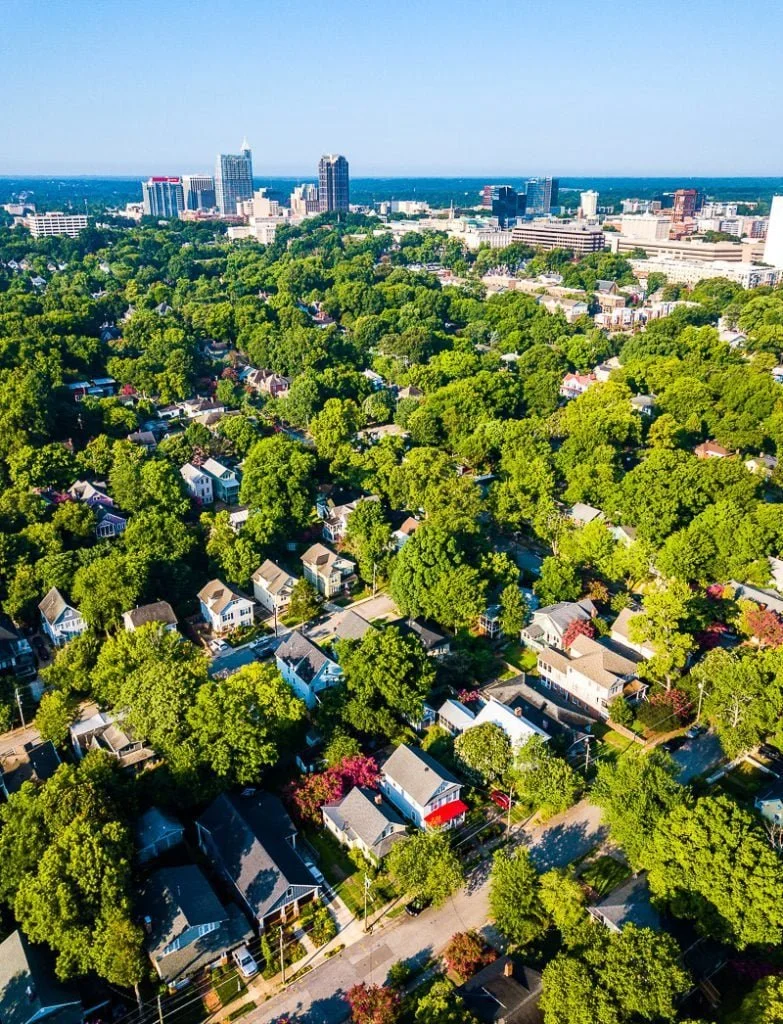 An aerial view of a Oakwood and Mordecai neighborhood with many houses surrounded by lush green trees, with a city skyline and taller buildings in the distance under a clear blue sky.