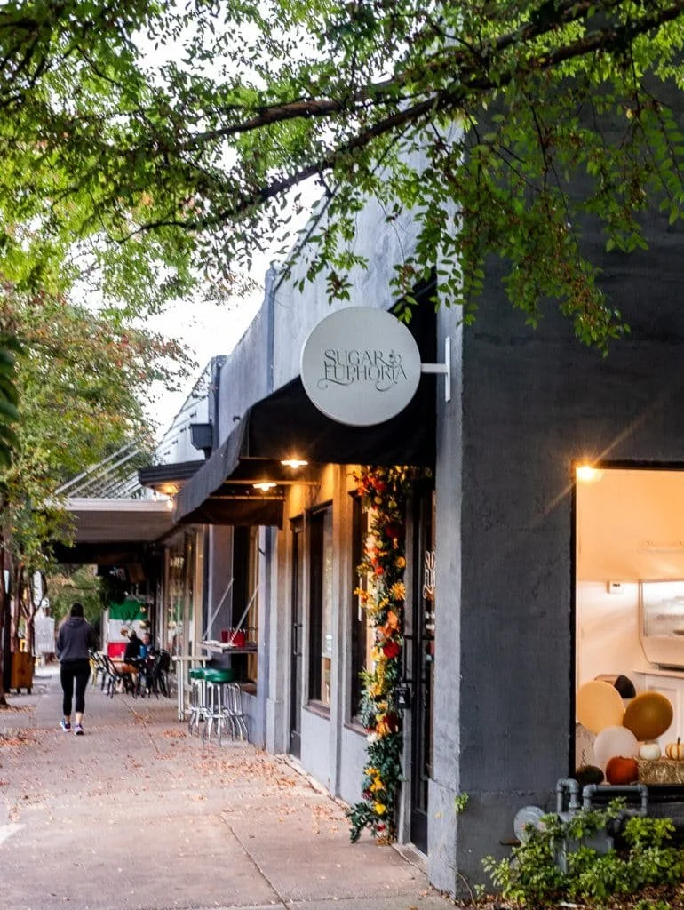 Street view showing Sugar Euphoria bakery with black awning and floral decoration outside. People walking and sitting at outdoor tables, trees lining the sidewalk.