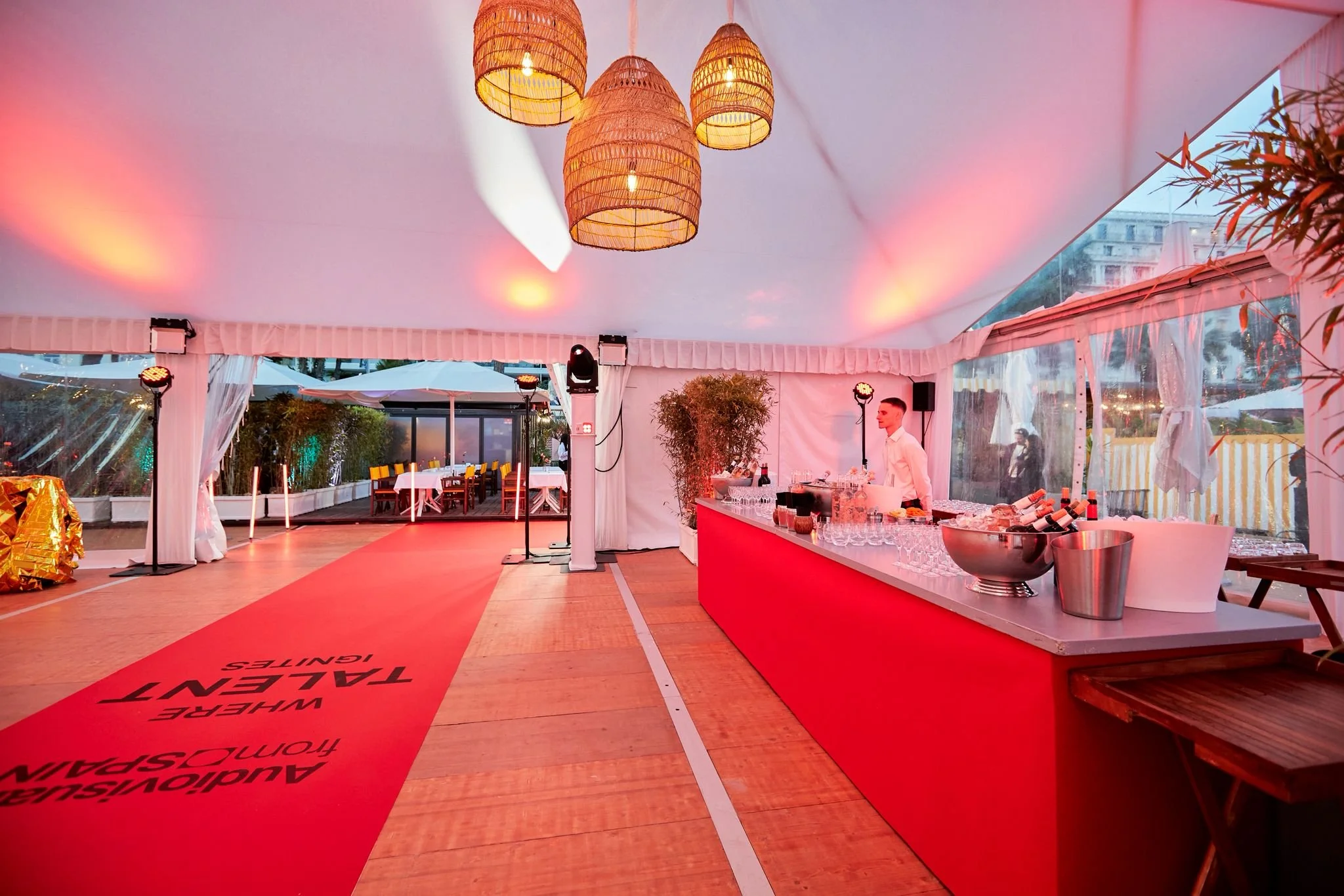Une tente lumineuse blanche avec un bar rouge à l'intérieur, comprenant des verres, des bouteilles de vin, et un barman. Il y a aussi des tables avec parasols à l'extérieur, et un éclairage chaud avec des lustres en osier suspendus.