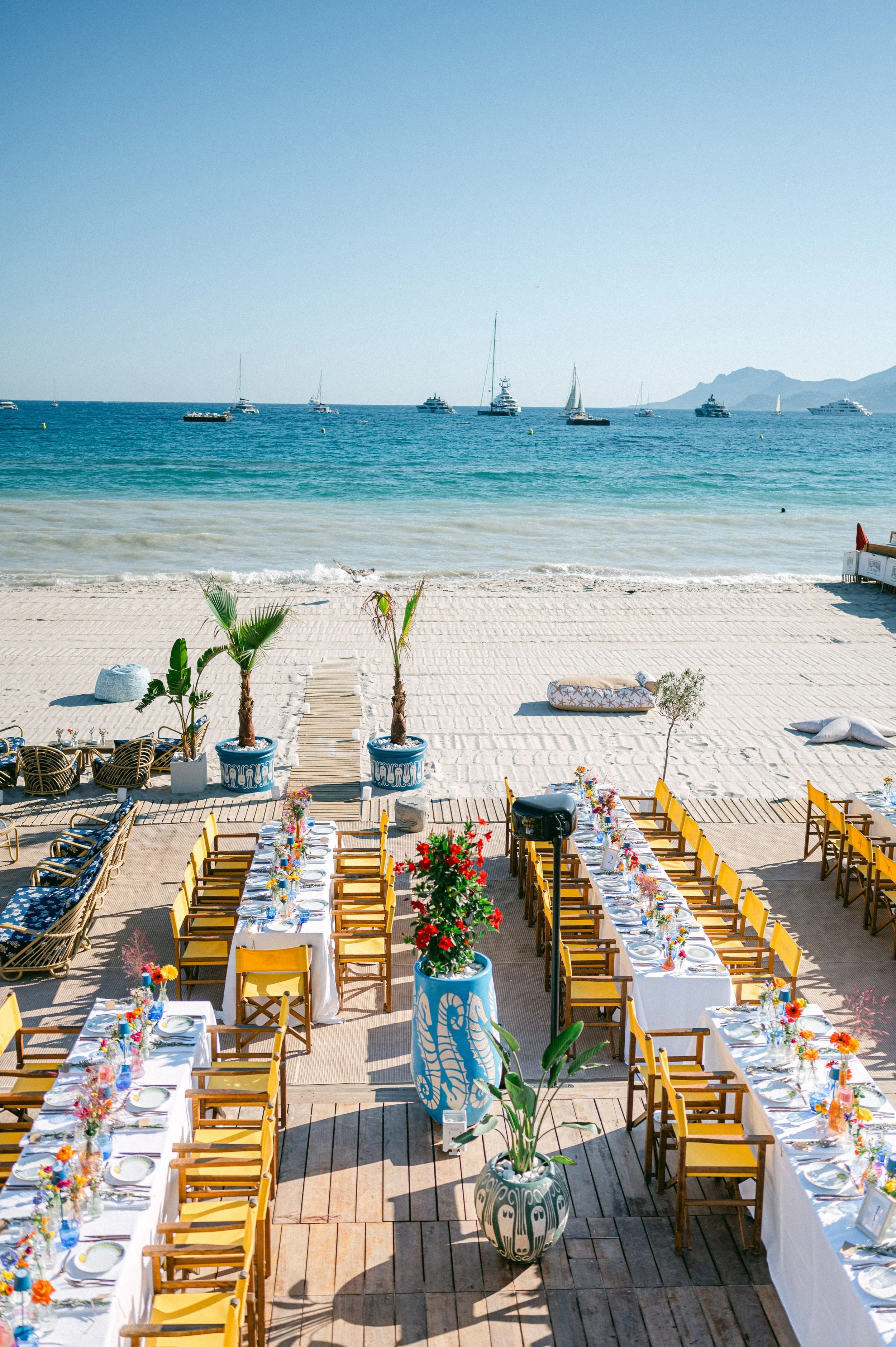 Une salle en plein air avec une table longue et plusieurs chaises jaunes, décorée de fleurs, face à la mer avec des yachts et des montagnes à l'horizon, avec des plantes vertes en pots