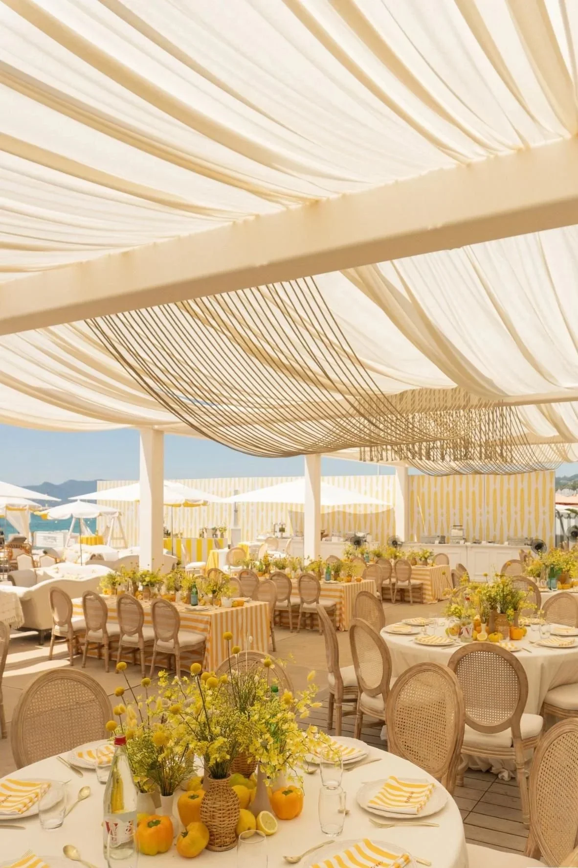 Une scène de fête en plein air avec des tables décorées de fleurs jaunes, citrons et poivrons, sous un grand canopy blanc avec une décoration de rubans noirs et blancs, dans un cadre ensoleillé.