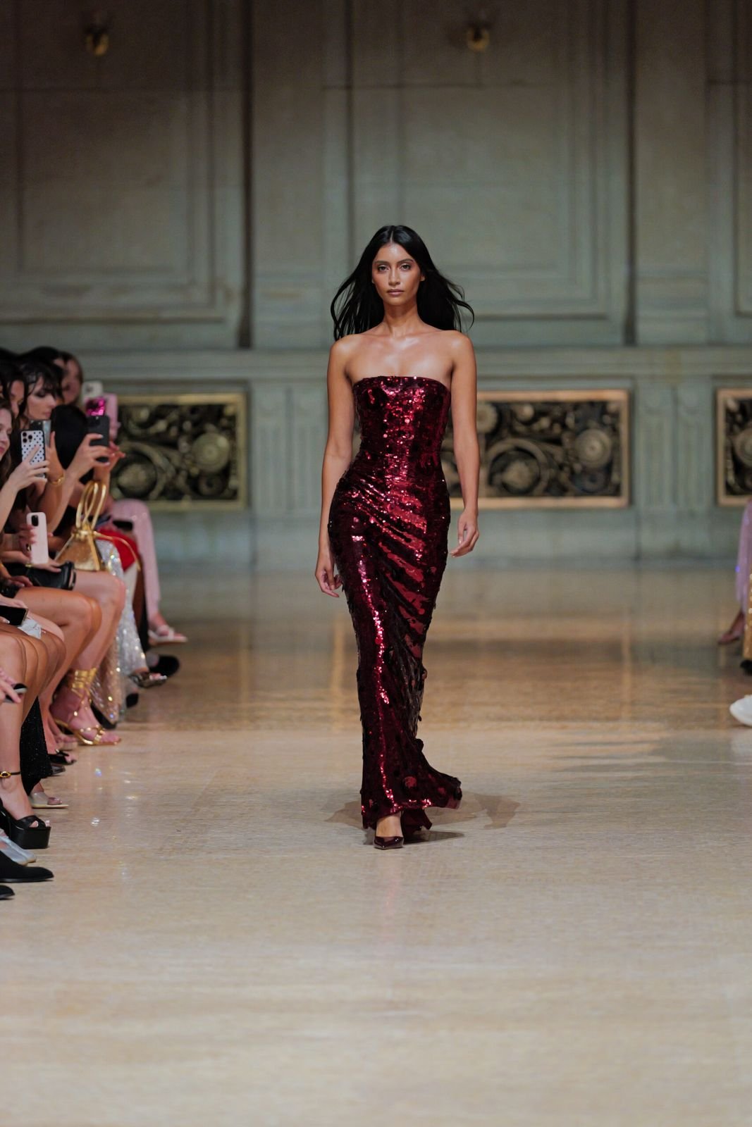 Une femme marche sur un podium de mode, portant une robe longue en sequins rouges, lors d'un défilé.