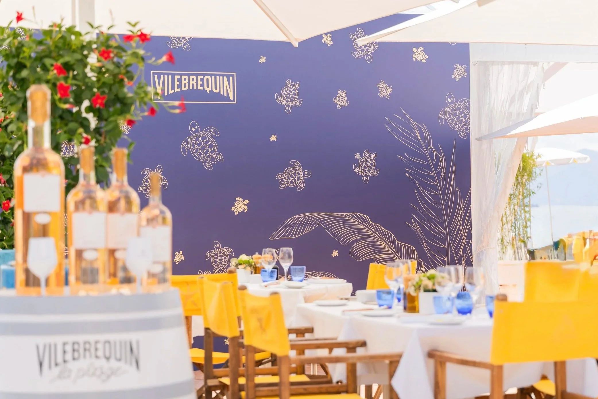 Décor d'événement en plein air avec un mur bleu orné de dessins de tortues et de feuilles, tables dressées avec des nappes blanches et des chaises jaunes, bouteilles d'alcool et verres sur la table.