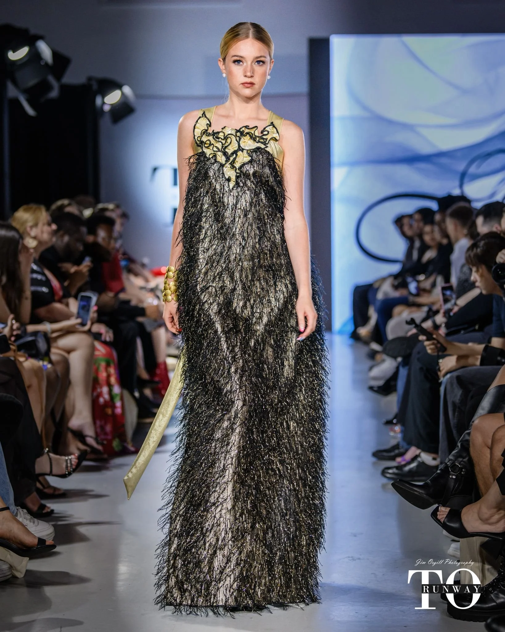 Une femme marche sur un podium lors d'un défilé de mode. Elle porte une robe longue en tissu noir et beige avec des détails en paillettes et un motif en relief. L'audience regarde et prend des photos, la scène est bien éclairée.