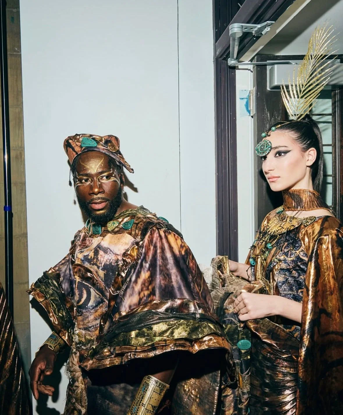 Deux modèles portent des vêtements et accessoires aux motifs or et animaux, dont un porte un couvre-chef décoratif et l'autre un bijou au front, dans un décor intérieur.