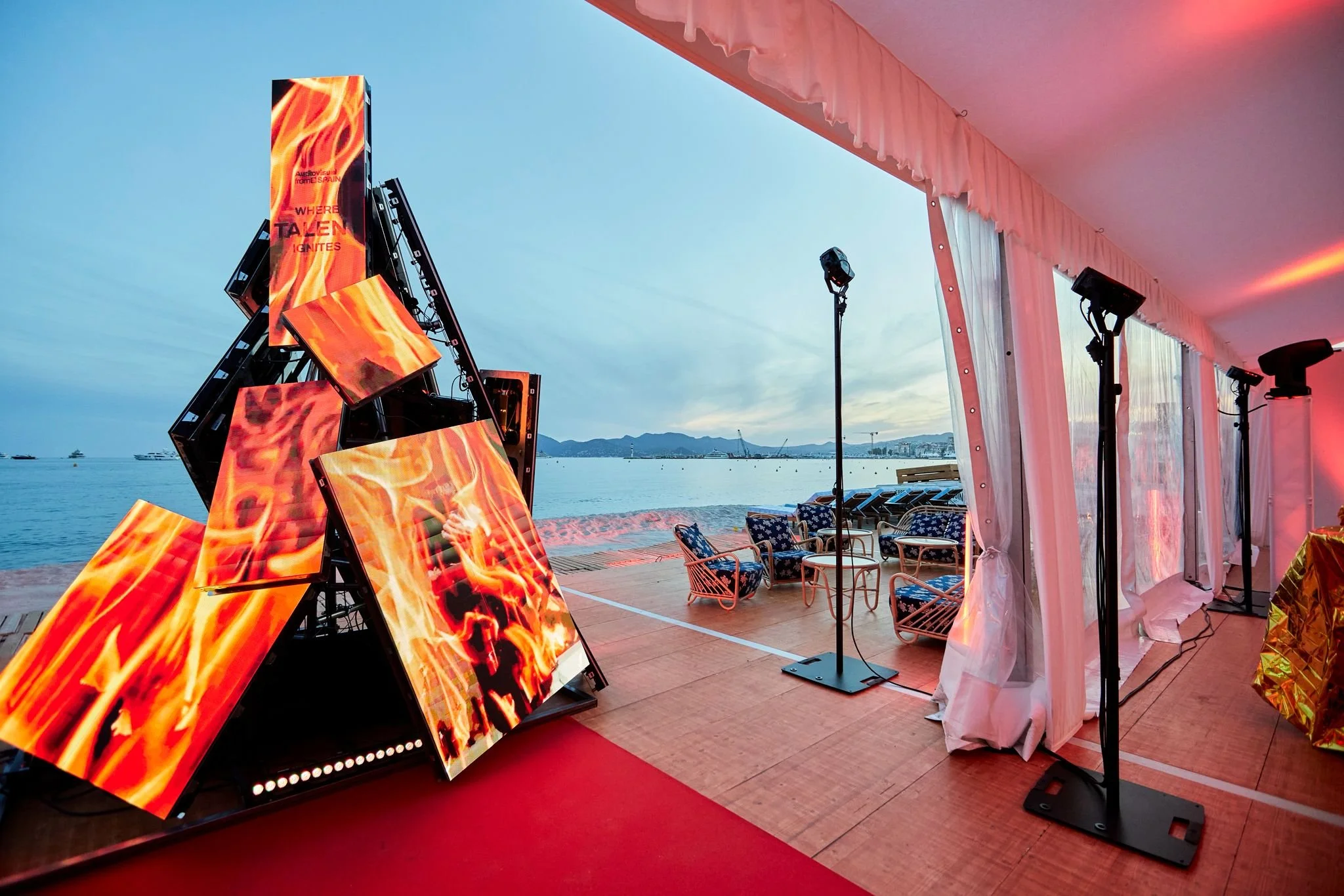 Installation d'art numérique avec plusieurs écrans affichant des flammes, à côté de la mer avec des chaises et une tente de fête en arrière-plan.