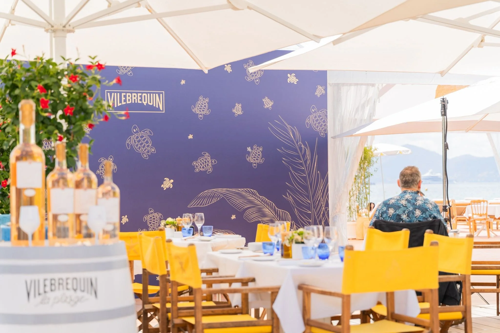 Vue d'un espace de repas en plein air avec des tables décorées de nappes blanches, des verres et des bouteilles de vin, entouré de chaises jaunes. En arrière-plan, un mur bleu avec des illustrations de tortues de mer et des feuilles, et des parasols blancs au-dessus. L'atmosphère est légère et ensoleillée, évoquant une célébration ou un événement festif près de la mer.