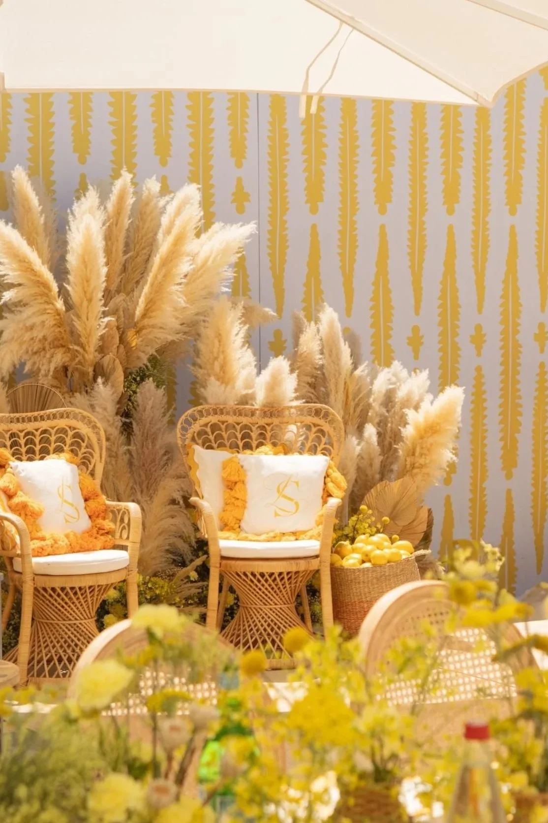 Décoration intérieure avec des chaises en rotin, coussins blancs ornés d'un monogramme doré, et des plantes de pampa jaune clair sur un fond jaune et blanc.