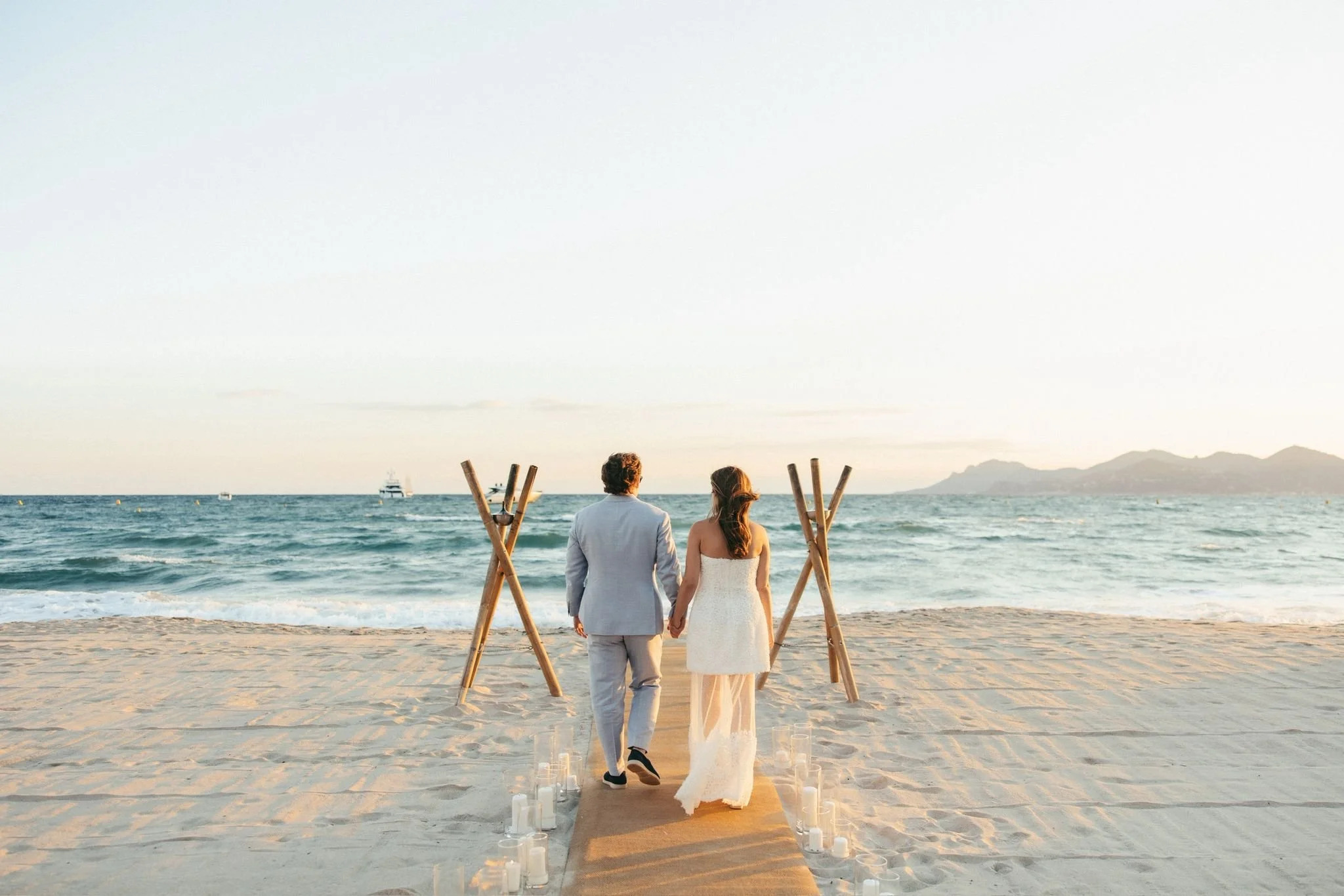 Un couple habillé en mariage marche sur la plage vers la mer, avec un chemin de bougies et un cadre en bois pour une cérémonie romantique au coucher du soleil.