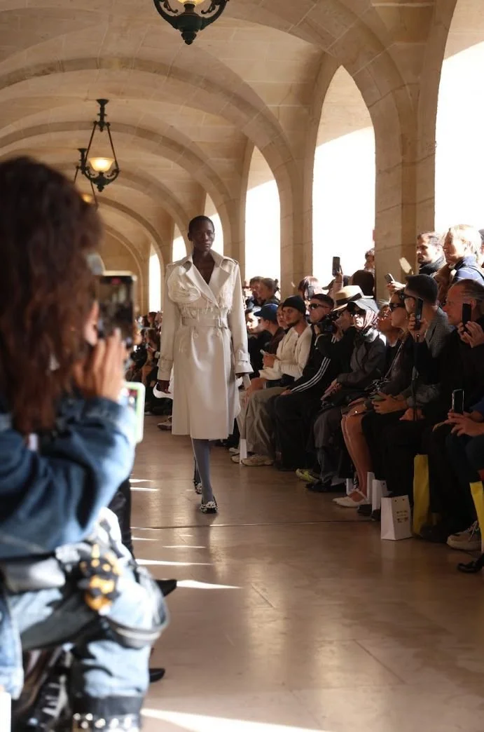 Un défilé de mode avec un modèle marchant sur le podium, entouré d'un public assis et pris en photo, dans une salle aux arches en pierre et aux lumières suspendues.
