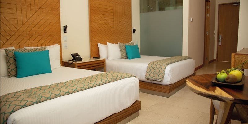 Hotel room with two double beds, pillows, and bed runners, nightstand with telephone, and a round wooden table with fruit.