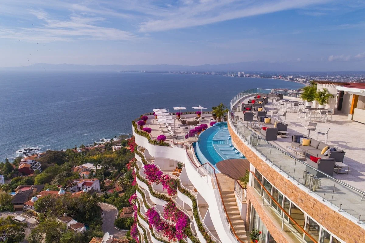 A luxury rooftop resort with a swimming pool, lounge area, and seating overlooking the ocean and cityscape.