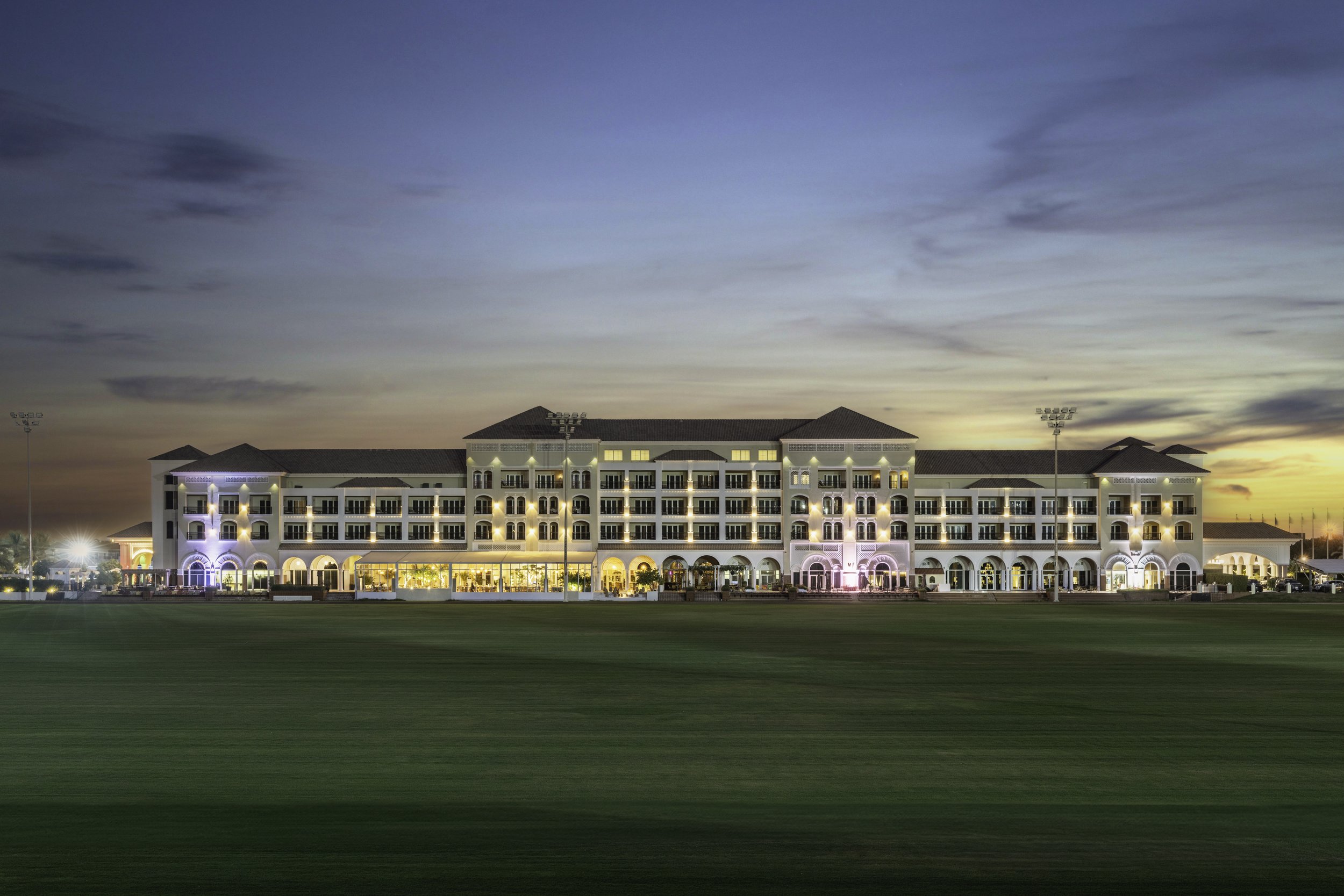 A large hotel building at dusk with colorful lights illuminating the facade, surrounded by a green lawn and tennis courts, under a sky with some clouds.