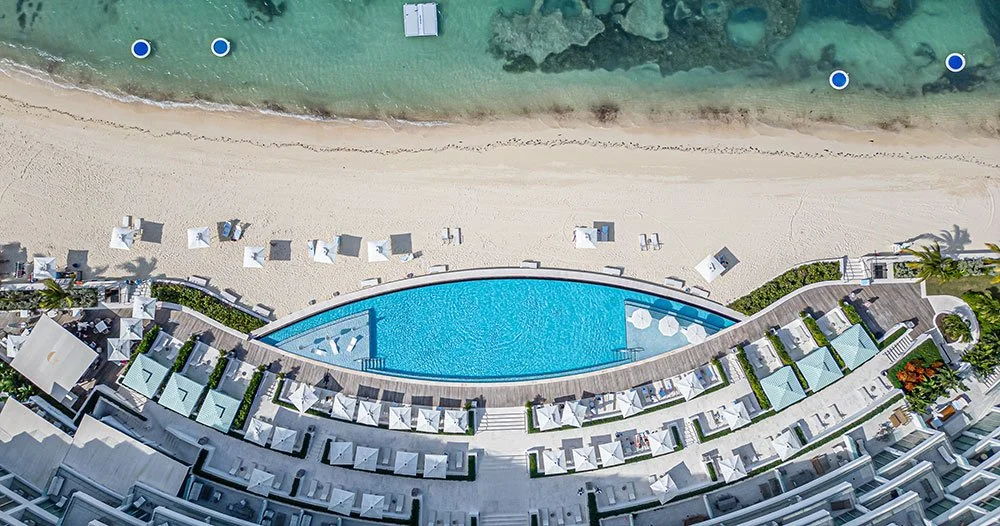Aerial view of a beach with a swimming pool surrounded by hotel rooms, beachfront umbrellas, and lounge chairs, near the ocean.