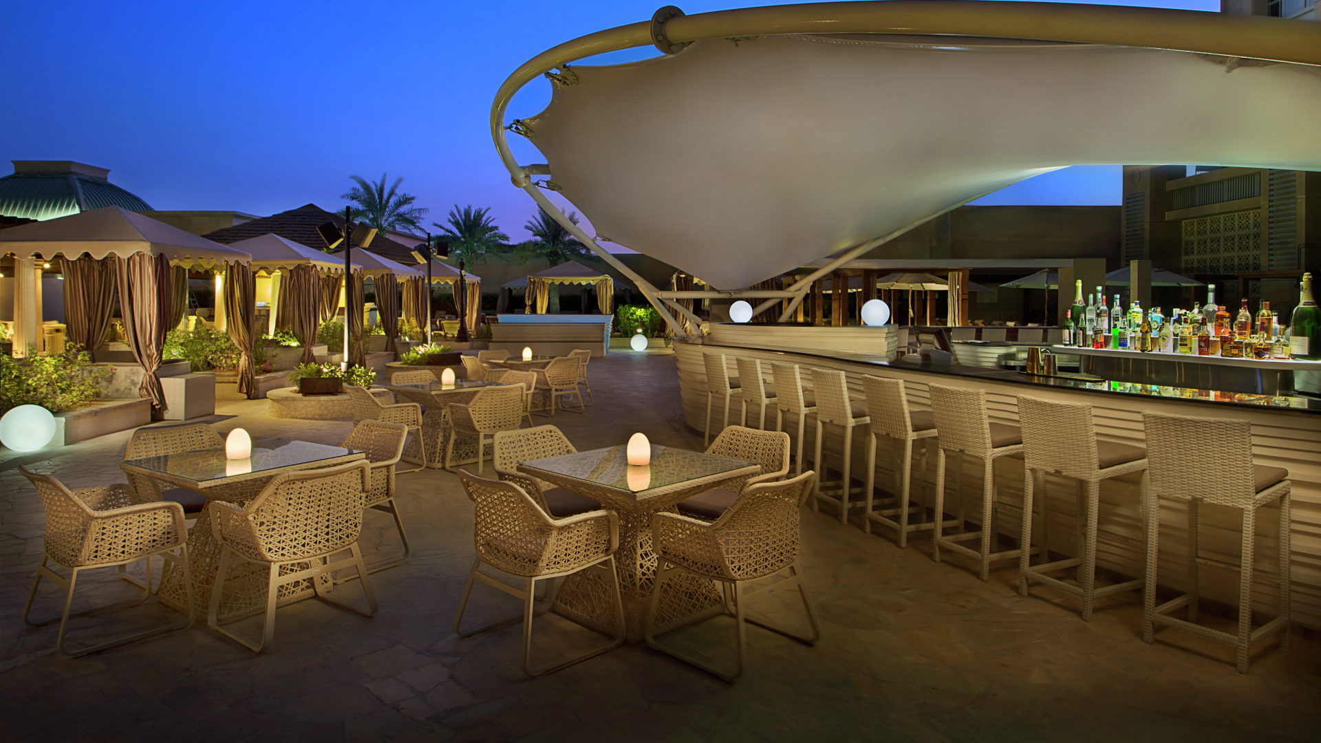 Outdoor lounge area at dusk with tables, chairs, bar, and string lights, under a large overhang