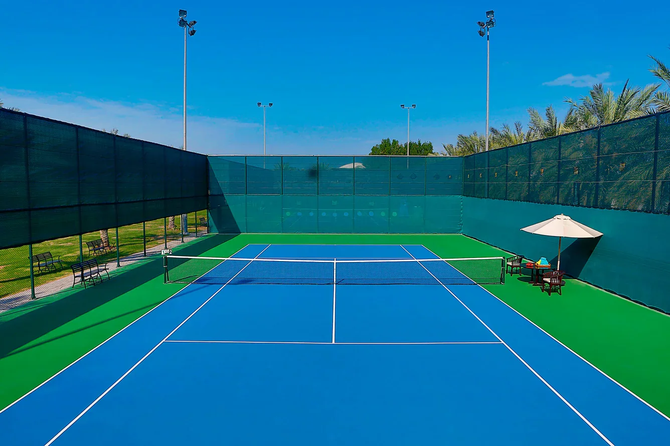 Empty blue tennis court with surrounding green area, benches, and a table with an umbrella, enclosed by a high fence under a clear blue sky.