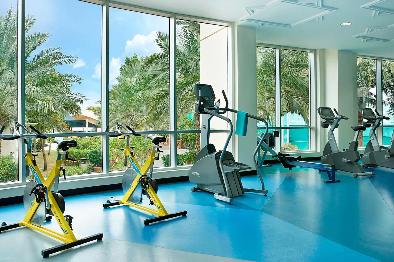 Indoor gym with stationary bikes, elliptical machines, and treadmills near large windows showing green trees and blue sky outside.