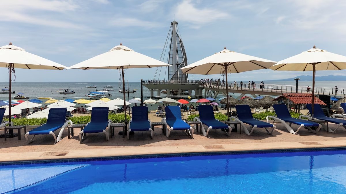 Poolside view at a beach resort with blue lounge chairs and white umbrellas in the foreground, sandy beach with colorful umbrellas and a pier with a sail-shaped structure in the background, and boats on the water.