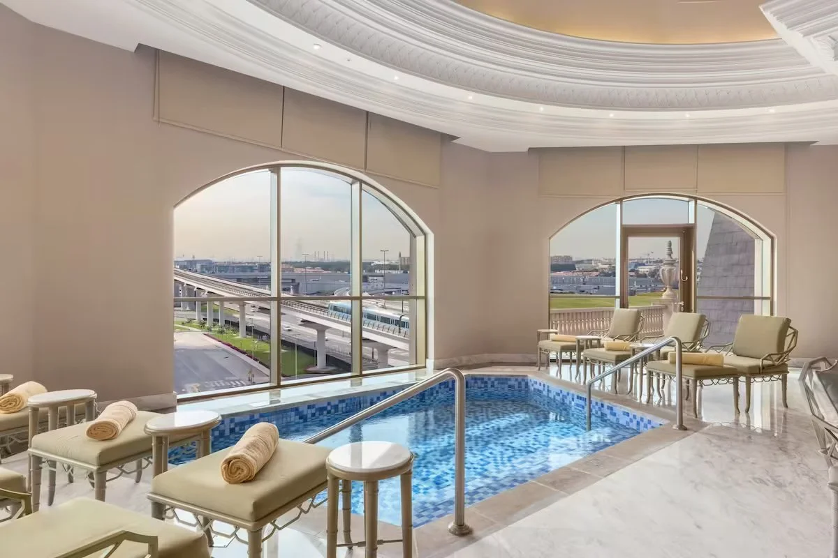Indoor pool area with large arched windows showing an outdoor view of a train station and cityscape, lounge chairs with rolled towels, and marble flooring.