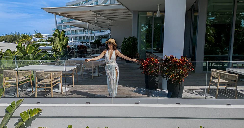 Woman in a wide-brimmed hat and white dress standing on a balcony with glass railing, surrounded by outdoor furniture and potted plants, with a building and greenery in the background.