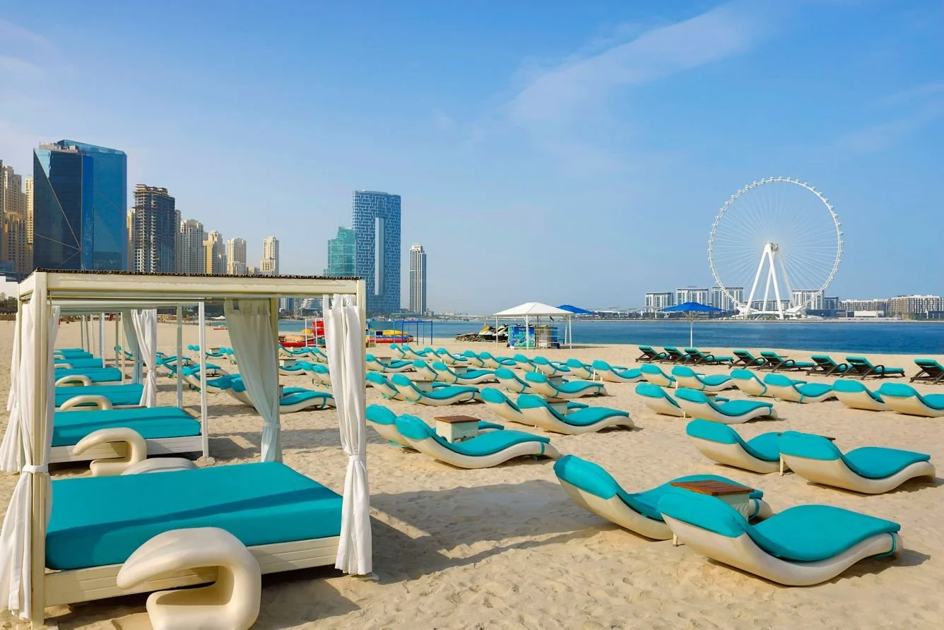 Empty beach loungers and cabanas on sandy shore with city skyline and Ferris wheel in the background under a clear blue sky.