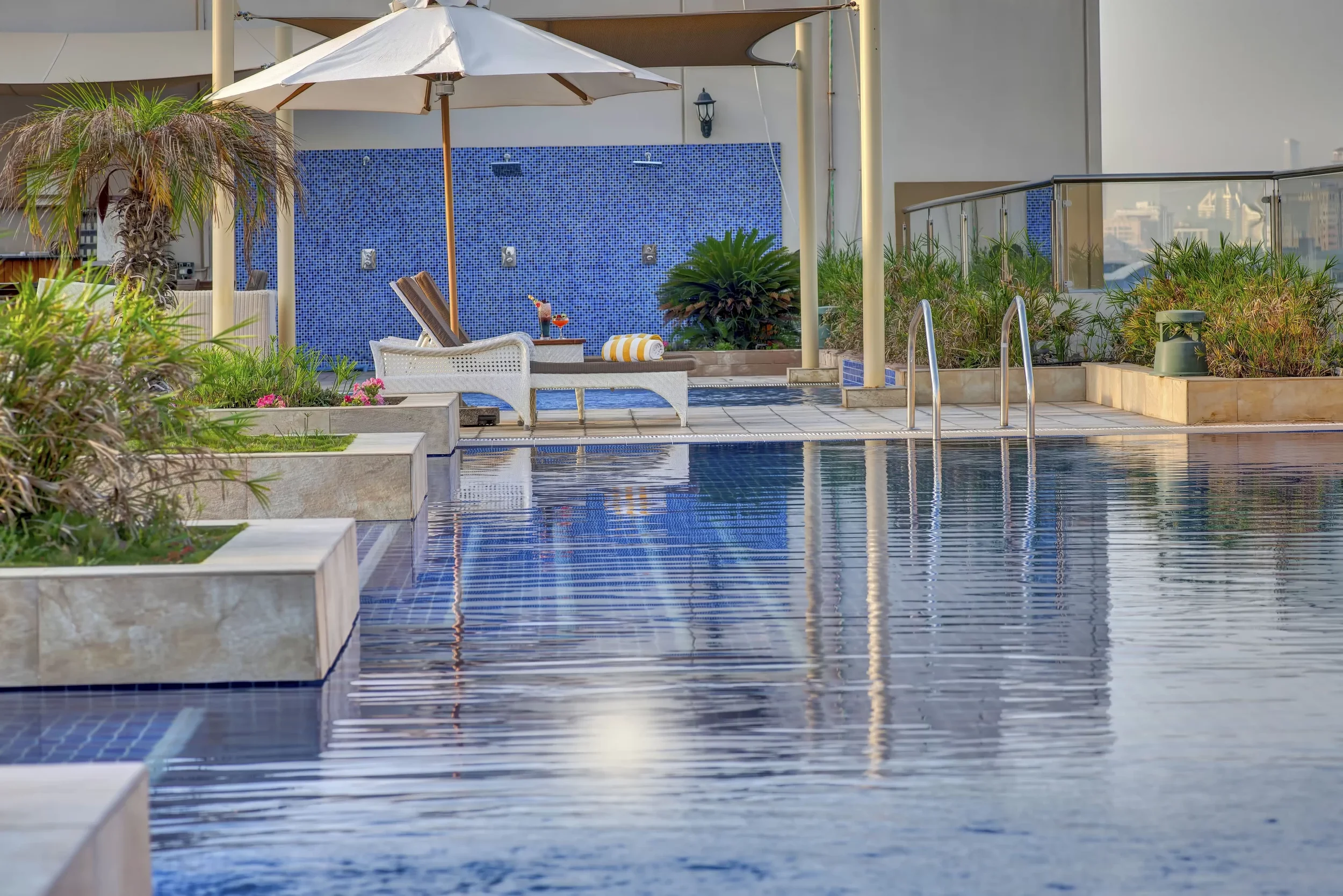 A rooftop swimming pool area with lounge chairs, a white parasol, lush plants, and a blue mosaic wall with outdoor showerheads.