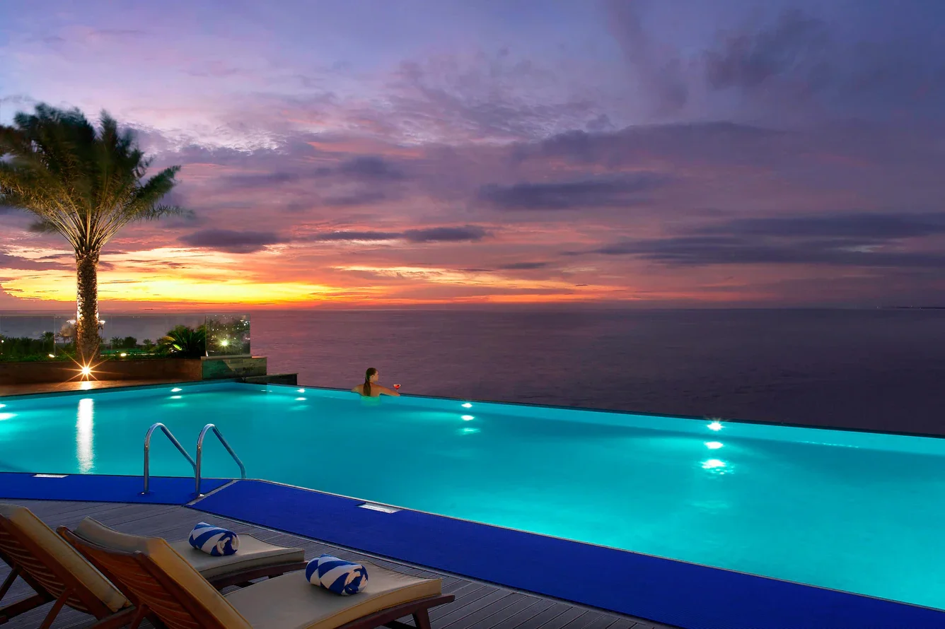 A person relaxing in a pool with an ocean view during sunset, with a palm tree and lounge chairs nearby.