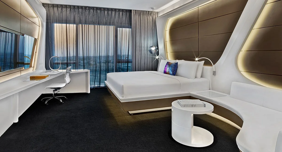 Modern hotel room with a large bed, a desk with a chair, a flat-screen TV, and large windows with curtains showing an urban city view.