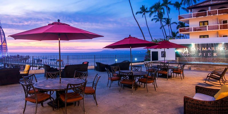 Outdoor patio with tables, chairs, and large red umbrellas overlooking the ocean at sunset, with palm trees and a building with a sign that reads 'San Marino Pier'.
