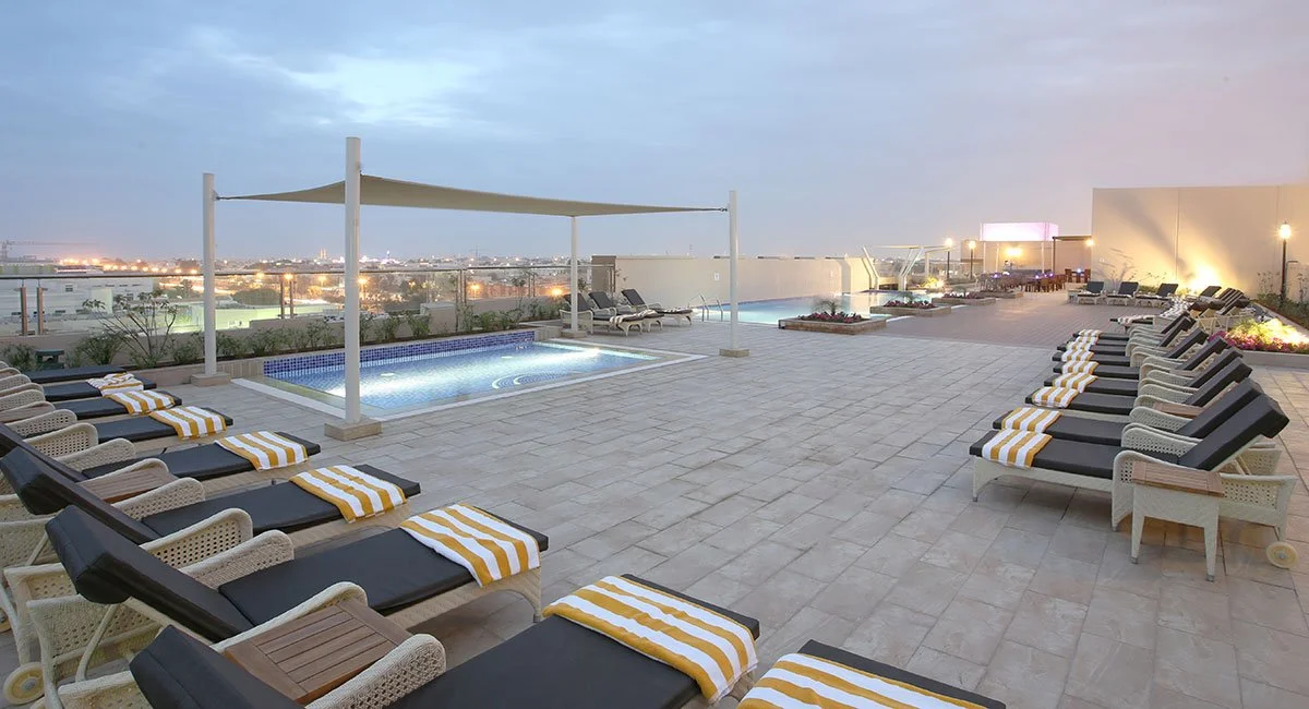Rooftop pool area with lounge chairs, a hot tub, and city skyline view at sunset.