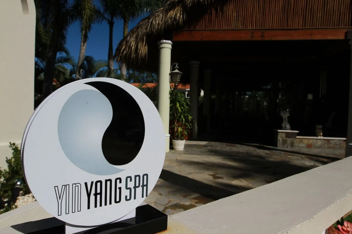 Sign for Yim Yang Spa with a black and white yin-yang symbol outside a tropical resort with palm trees and a thatched roof structure.