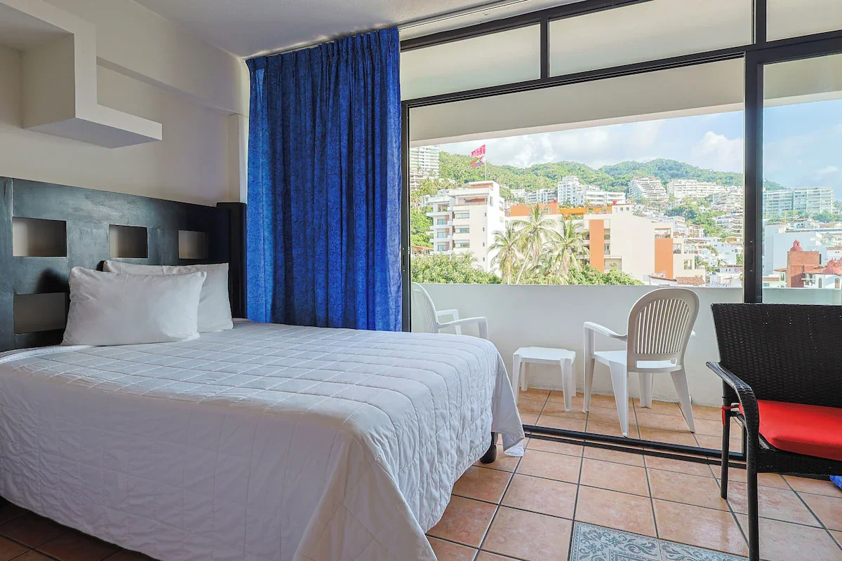Hotel room with a bed, blue curtains, and a balcony with outdoor chairs overlooking a city with colorful buildings and greenery.