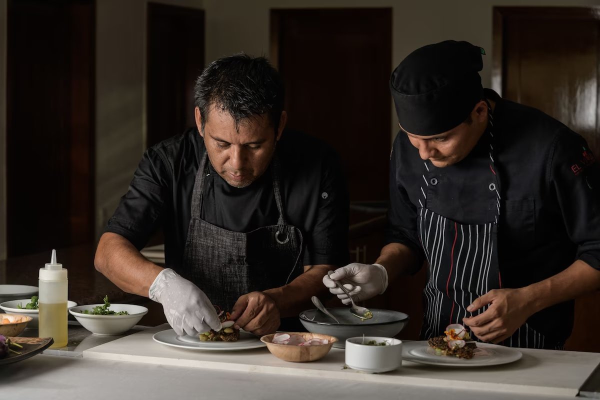 Two chefs in black uniforms and aprons preparing gourmet dishes in a professional kitchen.