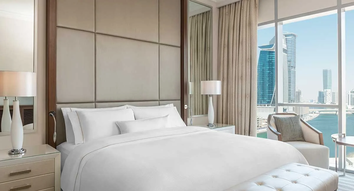 A luxury hotel room with a large white bed, bedside lamps, a sitting chair, and a window with a view of a city skyline including a tall modern building and a river.
