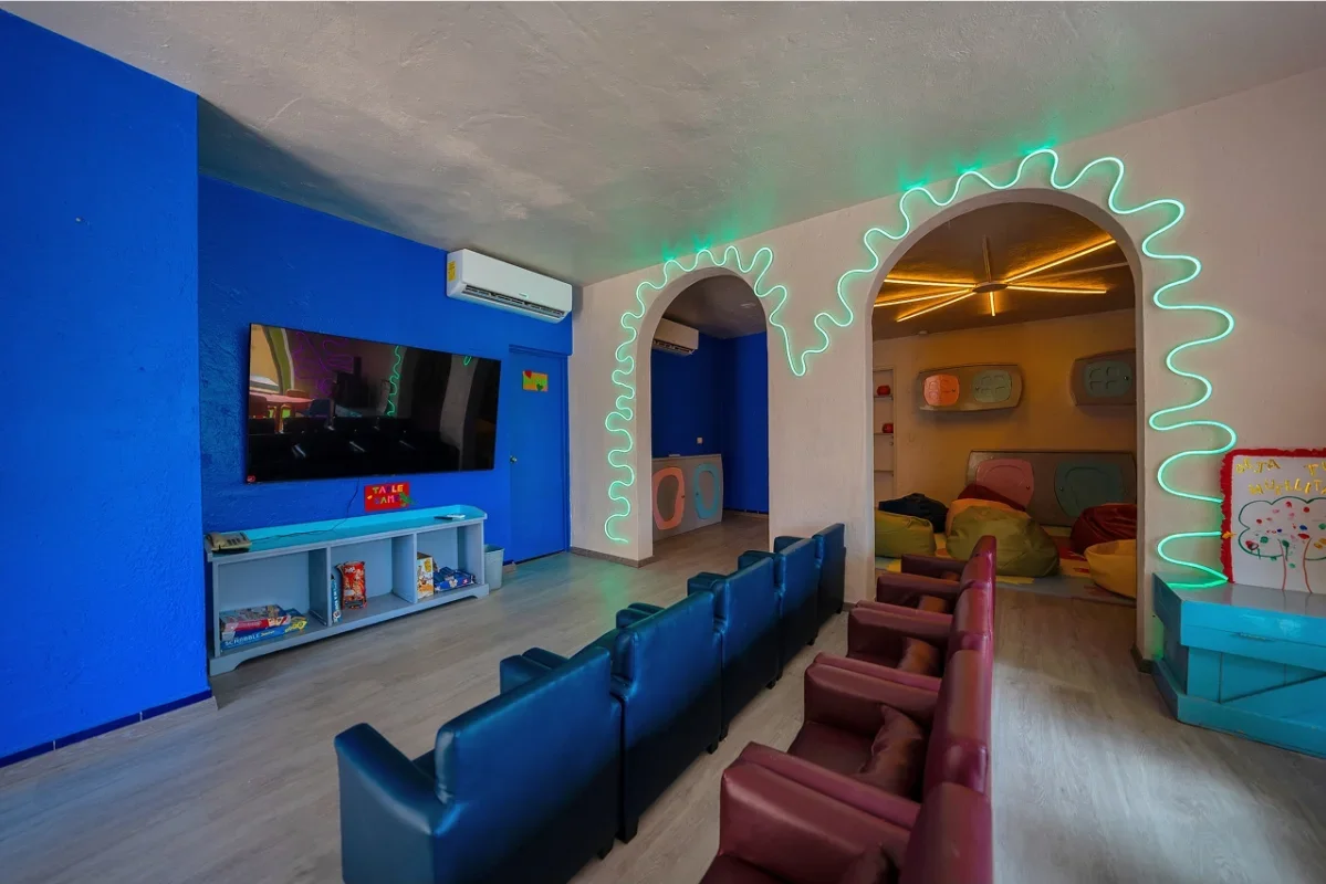 Colorful lounge with a blue accent wall, flat-screen TV, and gaming consoles, featuring a mix of blue and maroon chairs, decorative neon lights around archways, and a cozy area with cushions and soft lighting.