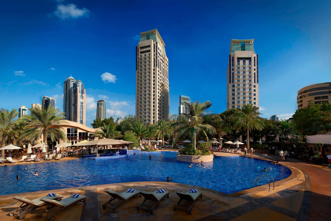Swimming pool area with lounge chairs, umbrellas, and palm trees in front of tall city buildings on a sunny day.