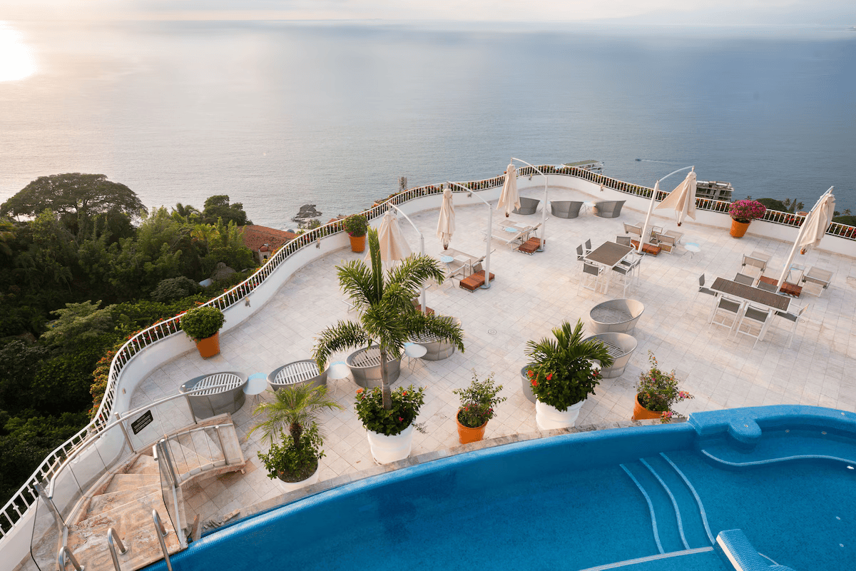 An empty outdoor terrace with lounge chairs, tables, umbrellas, potted plants, and a view of the ocean.