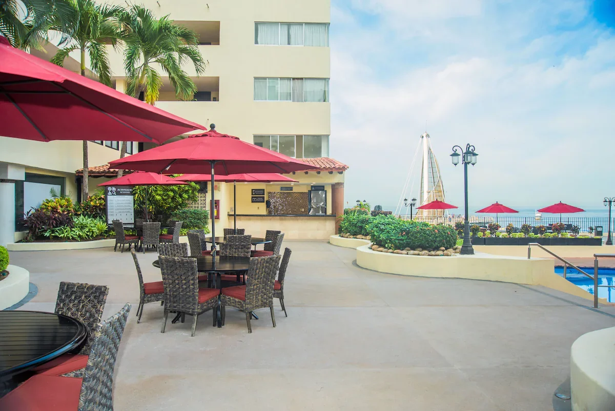 Outdoor patio area with red umbrellas, wicker chairs and tables, some with red cushions, around a beige building with palm trees, overlooking a marina with sailboats and an ocean view on a partly cloudy day.