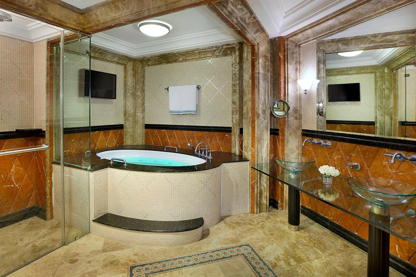 Luxury bathroom with a large bathtub, double glass sinks, mirror, wall-mounted TV, and marble walls and floors.