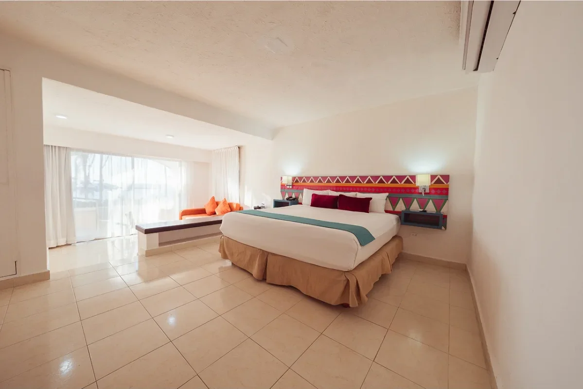 Hotel room with a large bed, colorful headboard, nightstands with lamps, and a sitting area with orange cushions near a large window with sheer curtains.